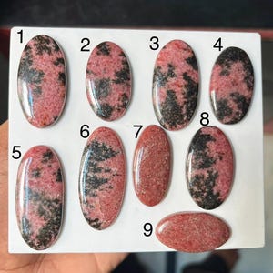 Puede incluir: Nueve cabujones de rodonita de forma ovalada dispuestos sobre una superficie blanca, numerados del uno al nueve. Cada piedra muestra un patr&oacute;n &uacute;nico de inclusiones minerales rosas y negras, con superficies pulidas lisas y bordes redondeados.