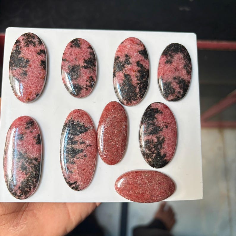 Puede incluir: Una colecci&oacute;n de ocho cabujones ovalados pulidos con un patr&oacute;n moteado de inclusiones minerales rosas y negras. Estas piedras lisas y redondeadas muestran concentraciones variables de vetas oscuras sobre un fondo texturizado de color rosa polvoriento.