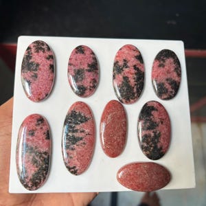 Puede incluir: Una colecci&oacute;n de ocho cabujones ovalados pulidos con un patr&oacute;n moteado en rosa y negro. Estas piedras lisas y redondeadas muestran concentraciones variadas de inclusiones oscuras sobre una base rosa vibrante, dispuestas sobre una superficie blanca.