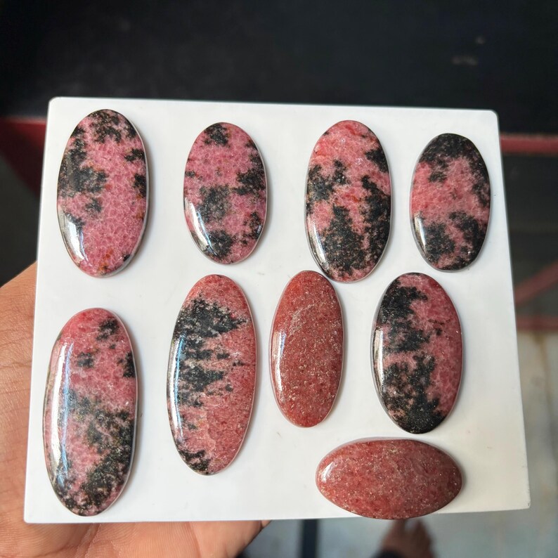 Puede incluir: Una colecci&oacute;n de ocho cabujones ovalados pulidos con un patr&oacute;n moteado en rosa y negro. Estas piedras lisas y redondeadas muestran concentraciones variadas de inclusiones oscuras sobre una base rosa vibrante, dispuestas sobre una superficie blanca.