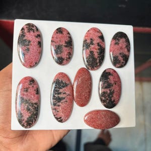 Puede incluir: Una colecci&oacute;n de ocho cabujones ovalados pulidos con un patr&oacute;n moteado de rosa empolvado e inclusiones minerales de color negro intenso. Estas piedras lisas y redondeadas est&aacute;n dispuestas sobre una superficie blanca plana.
