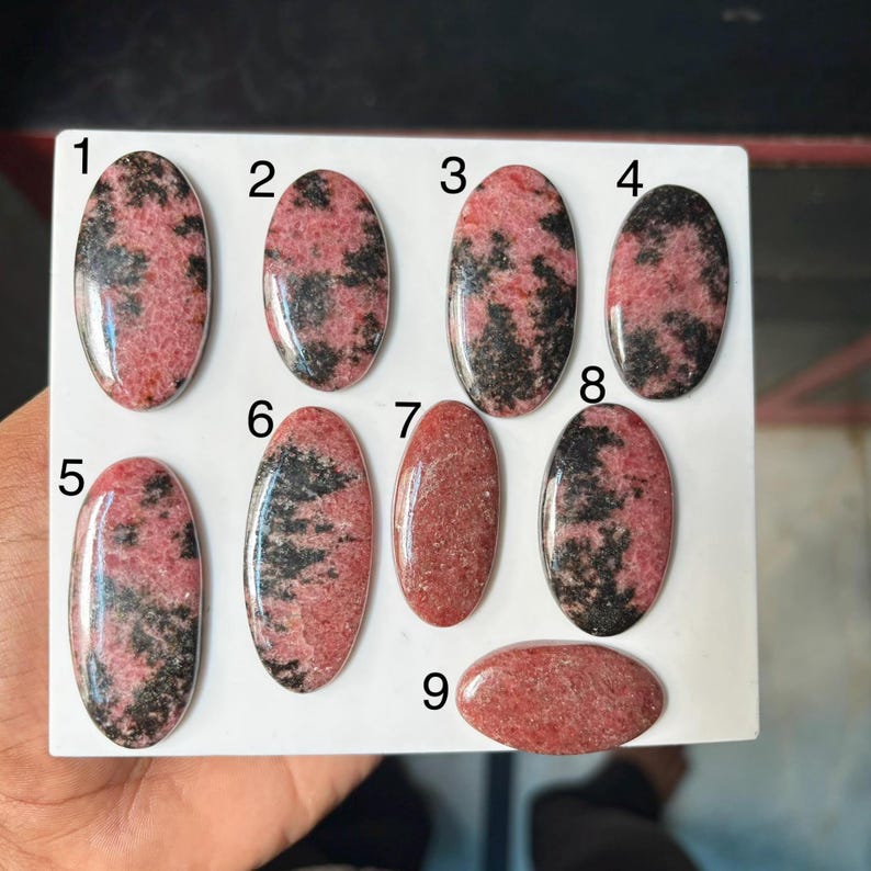 Puede incluir: Nueve cabujones de rodonita de forma ovalada dispuestos sobre una superficie blanca, numerados del uno al nueve. Cada piedra muestra un patr&oacute;n &uacute;nico de inclusiones minerales rosas y negras, con superficies pulidas lisas y bordes redondeados.
