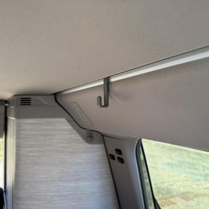 Pode incluir: Um gancho de metal cinza fixado ao teto de um veículo. O gancho é projetado para pendurar objetos. O teto é cinza claro, e a imagem mostra uma parte do interior, incluindo uma janela e uma cortina.