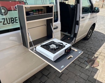 May include: A white portable camping stove with a black burner sits on a fold-down table attached to the side of a white van. The table is made of wood and has a black surface. The van has a black cabinet with shelves above the table.