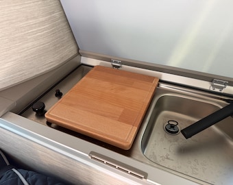 May include: A wooden cutting board rests on a stainless steel surface next to a sink. The cutting board is rectangular with a light brown color. The sink has a drain and a black faucet. The scene is part of a camper van kitchen.