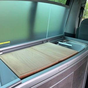 Op de afbeelding: Een witte bestelwagen met een ingebouwde keuken. Het aanrecht heeft een gootsteen, twee houten snijplanken en een grijze kast.