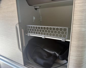 May include: A light gray, honeycomb-patterned shelf inside a cabinet. Below the shelf is a black bag. The cabinet has a light wood grain finish and a silver handle.