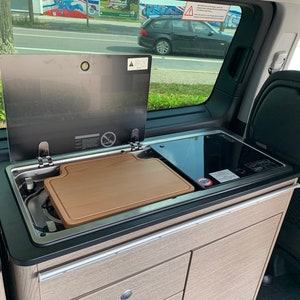 Op de afbeelding: Een zwart aanrecht met een ingebouwde gootsteen, een snijplank en een zwart elektrisch fornuis. Het aanrecht bevindt zich in een busje of camper.