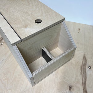 Puede incluir: Una caja de madera con una tapa con bisagras y un pequeño agujero en la parte superior. La caja tiene dos compartimentos en el interior, uno de los cuales está dividido en dos compartimentos más pequeños.