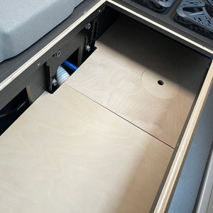 May include: Interior view of a camper van cabinet with a light wood paneling. The cabinet has a dark gray frame and includes a storage compartment, plumbing, and a section for a stove. The design is functional and space-efficient.