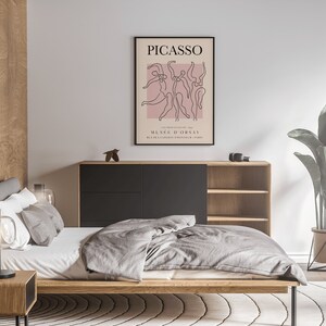 Puede incluir: Un interior de dormitorio con una estructura de cama de madera, un edred&oacute;n gris y una l&aacute;mpara negra sobre una mesita de noche de madera. Un p&oacute;ster enmarcado en negro con el texto "PICASSO" y un dibujo lineal de bailarines cuelga de la pared sobre una c&oacute;moda de madera con cajones negros. Una planta en maceta con hojas verdes se encuentra sobre un soporte de metal negro en una alfombra blanca.