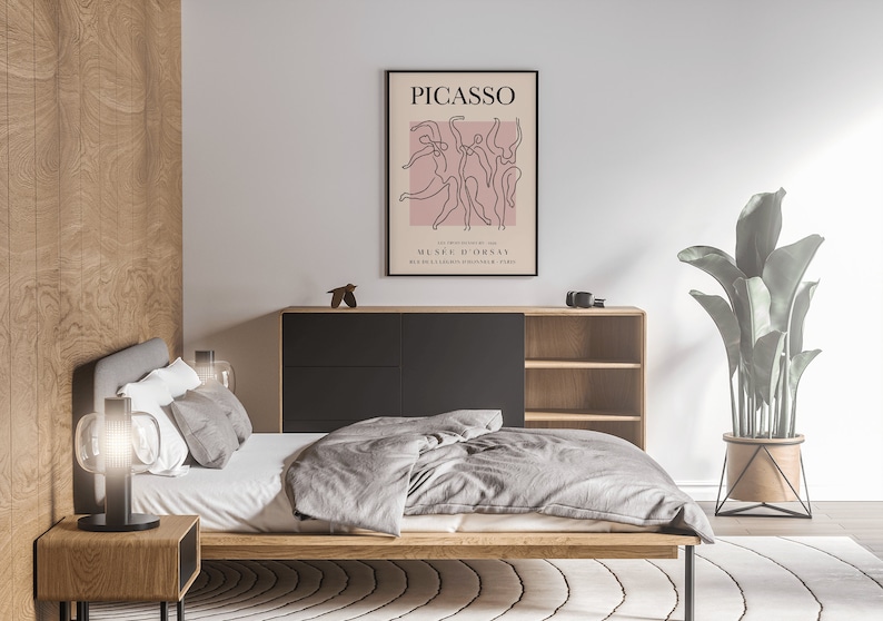 Puede incluir: Un interior de dormitorio con una estructura de cama de madera, un edred&oacute;n gris y una l&aacute;mpara negra sobre una mesita de noche de madera. Un p&oacute;ster enmarcado en negro con el texto "PICASSO" y un dibujo lineal de bailarines cuelga de la pared sobre una c&oacute;moda de madera con cajones negros. Una planta en maceta con hojas verdes se encuentra sobre un soporte de metal negro en una alfombra blanca.