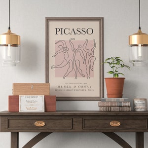 Puede incluir: Una impresi&oacute;n enmarcada de un dibujo de l&iacute;nea de bailarines con el texto "PICASSO" y "MUS&Eacute;E D'ORSAY" en un marco marr&oacute;n sobre una mesa de madera con dos l&aacute;mparas colgantes doradas y una pila de libros.
