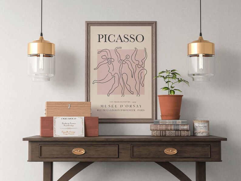 Puede incluir: Una impresi&oacute;n enmarcada de un dibujo de l&iacute;nea de bailarines con el texto "PICASSO" y "MUS&Eacute;E D'ORSAY" en un marco marr&oacute;n sobre una mesa de madera con dos l&aacute;mparas colgantes doradas y una pila de libros.