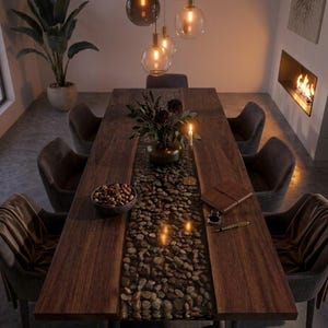 Pode incluir: Uma mesa de jantar retangular de madeira com um embutido central de pedras de rio lisas encapsuladas em resina transparente. A mesa é rodeada por oito cadeiras estofadas e contém uma tigela de nozes, um vaso, uma vela e um caderno.