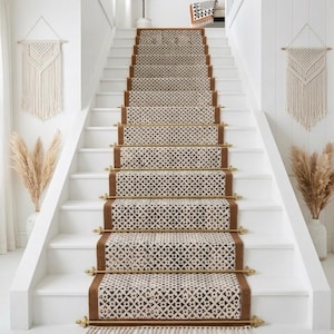 May include: A white staircase with a patterned runner rug. The rug has a brown border and a cream-colored geometric design. Decorative macrame wall hangings and pampas grass in vases flank the stairs. The stairs have brass rods.
