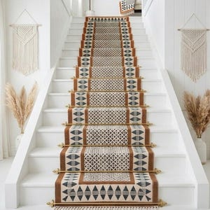 May include: Staircase runner with a geometric pattern in brown, black, and cream. The runner is installed on white stairs with brass rods. Decorative macrame wall hangings and pampas grass in vases flank the staircase.