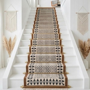 May include: A staircase with a runner rug featuring a geometric pattern in black and cream. The runner has a brown border and gold-colored rods. Macrame wall hangings and pampas grass in vases decorate the space.