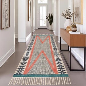 Peut inclure: Un tapis rectangulaire pour couloir avec un motif géométrique. Le tapis présente un fond gris clair avec un grand motif en forme de M de couleur corail au centre. Les bords sont bordés d'un motif triangulaire bleu marine et blanc, et d'une frange turquoise.