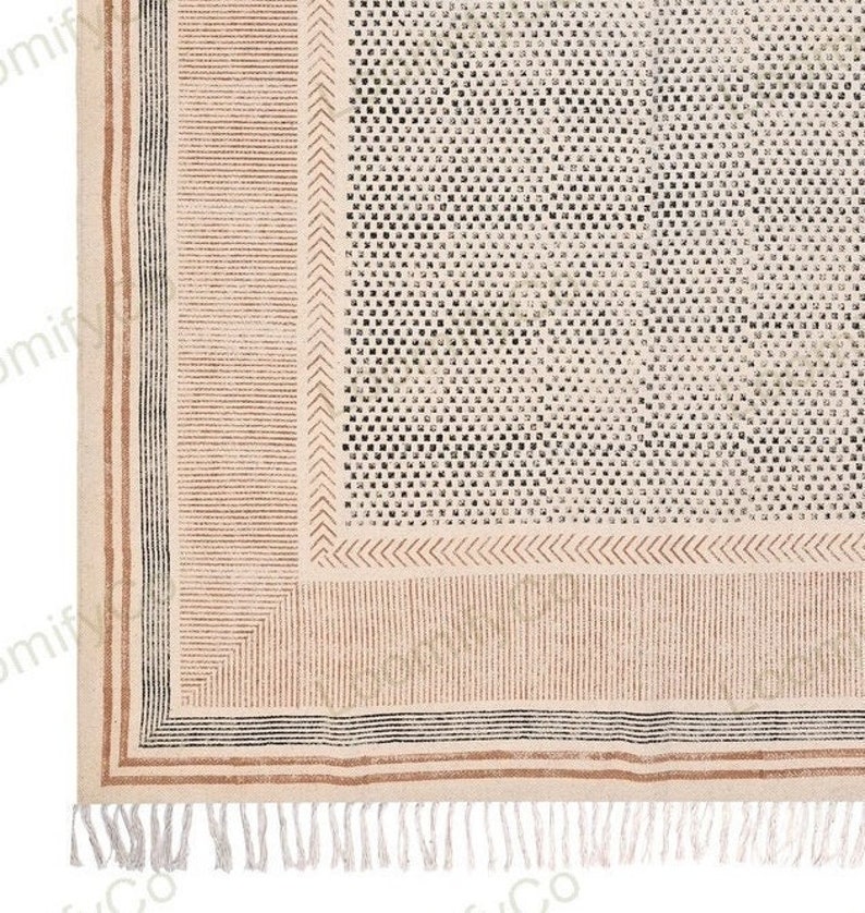 May include: A beige rug with a black and brown geometric pattern. The rug has a fringe border.