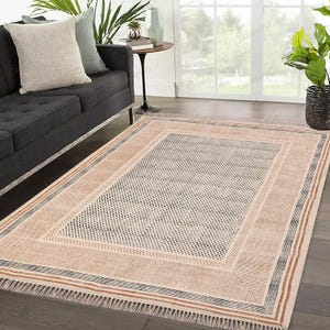 May include: A beige and brown patterned rug with a black and white geometric design. The rug has a fringe border and is laid out on a hardwood floor.