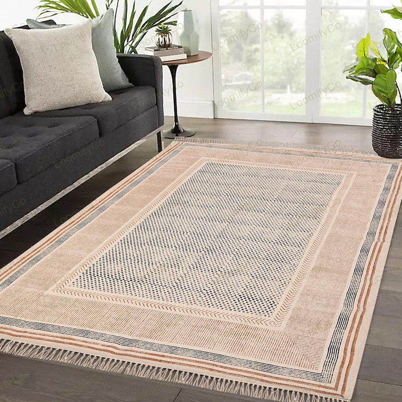 May include: A beige and brown patterned rug with a black and white geometric design. The rug has a fringe border and is laid out on a hardwood floor.