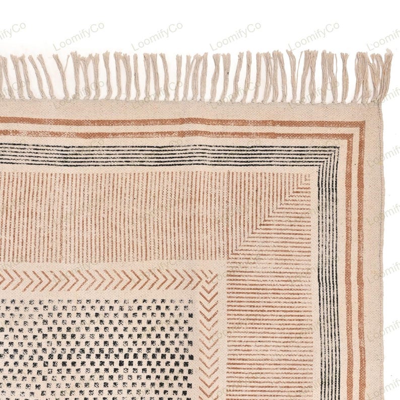 May include: A beige and brown patterned rug with a black and white geometric design in the center. The rug has a fringe border.