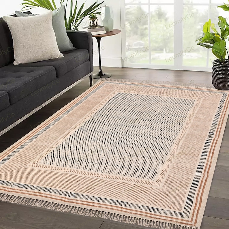 May include: A cream-colored rug with a black and brown geometric pattern. The rug has a fringe border and is laid out on a wooden floor.