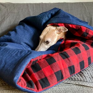 Puede incluir: Un perro está acurrucado dentro de una cama para perros de mezclilla azul y búfalo rojo y negro.