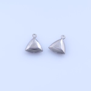 Puede incluir: Dos colgantes triangulares plateados con un pequeño lazo en la parte superior para sujetar a la joyería. Los colgantes tienen una superficie lisa y reflectante y una forma tridimensional. Se colocan sobre un fondo liso de color azul claro.