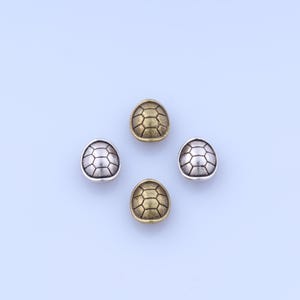 Peut inclure: Quatre perles en métal en forme de carapace de tortue. Deux sont de couleur argentée et deux de couleur dorée. Chaque perle a une surface texturée détaillée, ressemblant à une carapace de tortue. Les perles sont disposées sur un fond bleu clair.