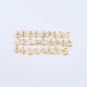 Puede incluir: Un conjunto de colgantes dorados con letras del alfabeto, de la A a la Z, sobre un fondo blanco. Las letras, con un diseño de burbujas redondeadas, son ideales para joyería o manualidades personalizadas.