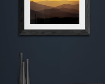May include: A framed photograph of a mountain range at sunset. The mountains are silhouetted against a bright orange sky. The frame is black and has a white mount.