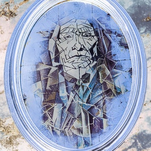 Puede incluir: Obra de arte ovalada enmarcada en plata, que representa a un hombre nativo americano en un estilo geométrico. La imagen utiliza una técnica cubista con tonos azules, grises y marrones. El marco es de color plata brillante.