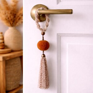 Może przedstawiać: Dekoracyjny wieszak na drzwi z plecioną beżową pętlą, okrągłą pomarańczową kulką filcową i długim beżowym frędzlem. Wieszak na drzwi jest przymocowany do mosiężnej klamki, dodając odrobinę stylu boho.