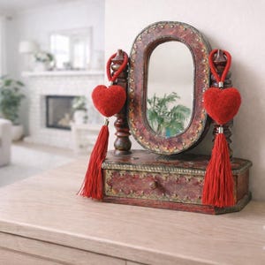 May include: A vintage dressing table mirror with an oval frame and a small drawer. The mirror is decorated with two red heart-shaped ornaments with long tassels. The wooden frame has a distressed, reddish-brown finish.