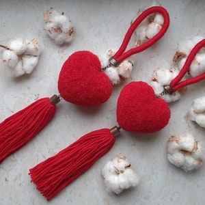 Peut inclure: Deux ornements rouges en forme de cœur avec des pompons et des cordons bouclés assortis. Les ornements sont entourés de boules de coton blanches et duveteuses. Les pompons et les cordons sont d'un rouge vif.