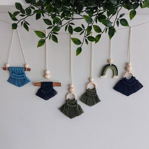 Peut inclure: Une guirlande de six tentures murales en macramé avec des perles et des chevilles en bois. Les tentures sont dans des tons de bleu et de vert, certaines avec des franges et d'autres avec un motif arc-en-ciel.