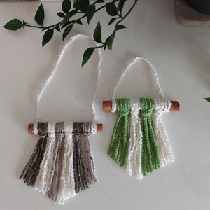 May include: Two handmade macrame wall hangings with cinnamon stick accents. One is white and beige with brown stripes, the other is white and green.