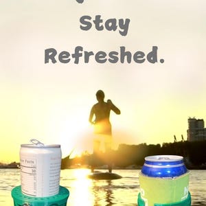 May include: Two green plastic cup holders with the text "cupsup" printed on them. The holders are holding aluminum cans. The cans are on a paddleboard in the water. The background is a sunset.