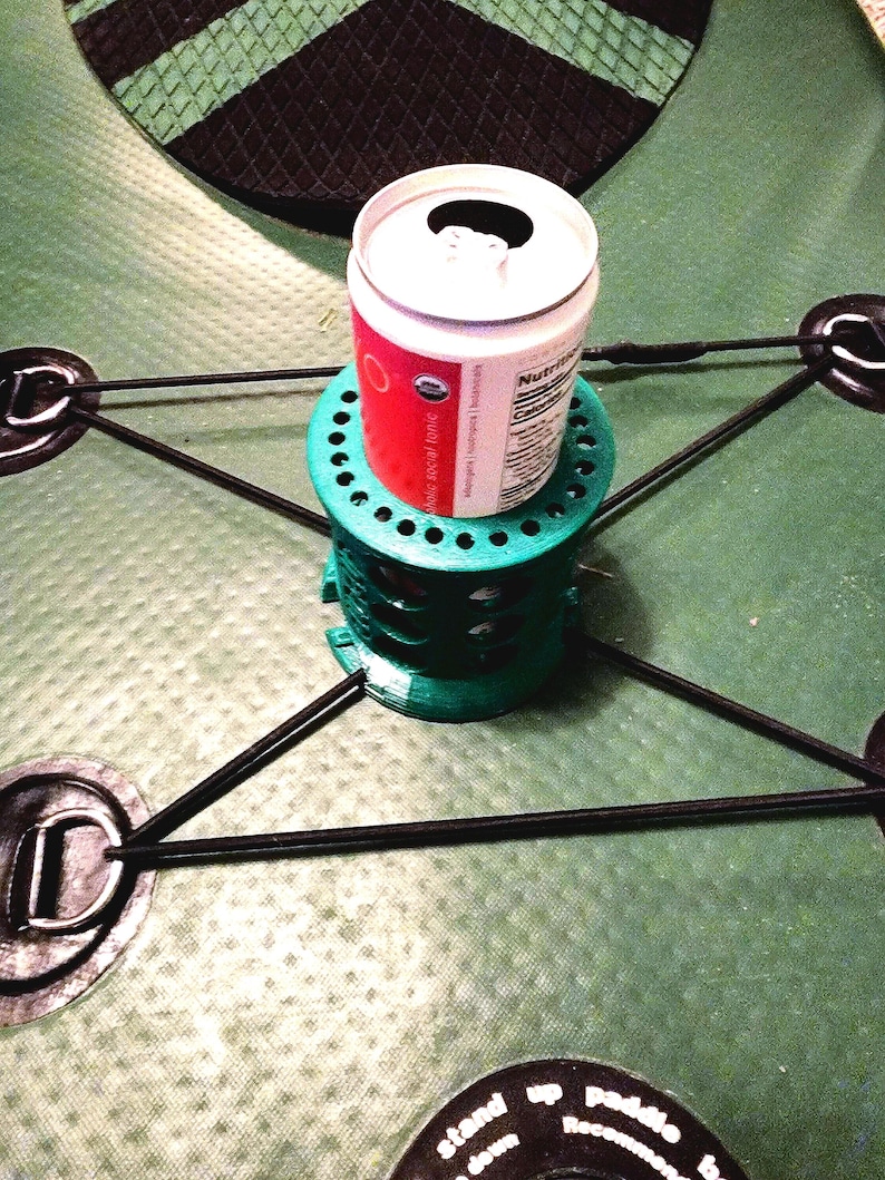 May include: A green 3D printed can holder with a black elastic strap attached to a green and black inflatable kayak. A can of soda is sitting on top of the holder.