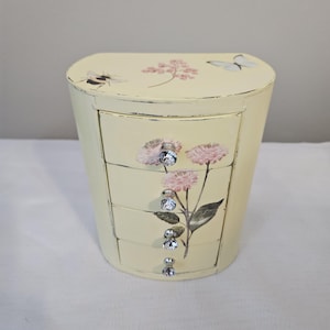 May include: A pale yellow jewelry box with three drawers and a curved top. The box is decorated with pink floral and butterfly designs. The drawers have crystal-like knobs. A bee is painted on the top.