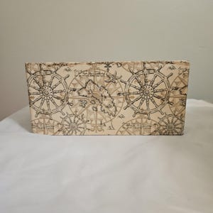 May include: Rectangular storage box with a nautical theme. The box is beige with a repeating pattern of ship wheels, compasses, and a map. The design is in shades of brown and tan.