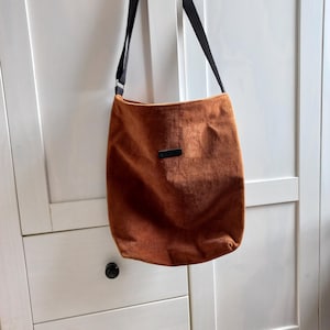Könnte beinhalten: Eine Umhängetasche aus orangefarbenem Cordstoff mit schwarzem Riemen. Die Tasche hat ein kleines, rechteckiges schwarzes Etikett. Das Design ist schlicht und praktisch, geeignet für den täglichen Gebrauch.