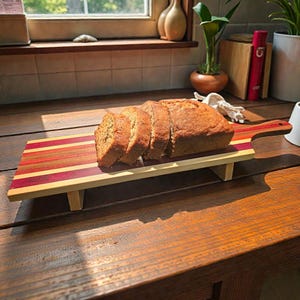 May include: A loaf of sliced bread rests on a wooden serving board with a handle. The board features a striped design in shades of red, purple, and yellow. The bread is light brown and appears freshly baked. A white mug sits nearby.
