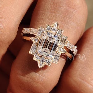 May include: A gold ring with a central, clear, rectangular gemstone, encircled by smaller, clear stones in a starburst pattern. The band is set with more clear, rectangular stones. The word "SKYRA" is visible on the ring.