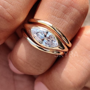 May include: A gold ring with a marquise-cut diamond center stone. The ring has a unique design with a double band and a polished finish. The word "SKYRA" is visible on the ring.