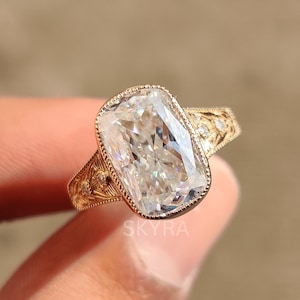 May include: A gold ring with a large, rectangular, clear gemstone. The ring has an ornate, engraved band and a beaded border around the gemstone. The word "SKYRA" is visible on the ring.