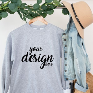 Può includere: Felpa grigia a maniche lunghe appesa a un appendiabiti di legno. La felpa ha la scritta "your design here" stampata in nero sul davanti.