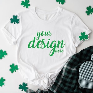 May include: White t-shirt with the text "your design here" in green on a white background. The t-shirt is surrounded by green shamrock stickers on a white surface.